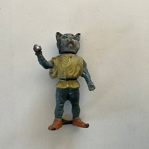 May include: A vintage figurine of a cat-like character in a medieval outfit. The figure has a gray cat head, a yellow tunic, and blue pants. It is holding a silver object in its raised hand. The figurine is approximately 4 inches tall.