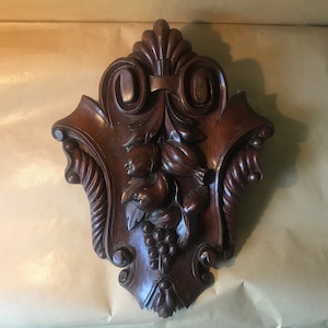Può includere: Una targa da parete in legno marrone scuro con un design intagliato di frutta e foglie. La targa ha la forma di uno scudo e ha un bordo decorativo.