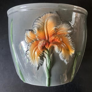 Peut inclure: Pot de fleurs en céramique grise avec un motif de fleur d'iris en relief. La fleur est orange et jaune avec des accents verts, sur un fond gris. Le pot a une finition lisse et brillante et une forme arrondie.