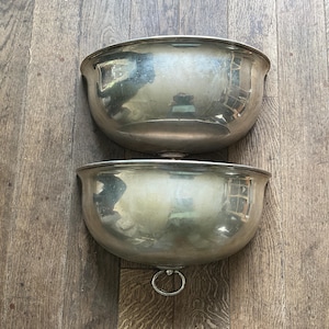 May include: Two antique silver-toned metal bowls. The bowls are semi-circular with a rounded rim and a small ring on the bottom. The bowls are reflective, showing the surrounding environment. The bowls are likely for decorative or serving purposes.