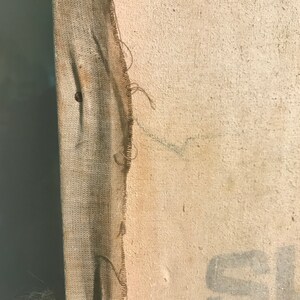 May include: Close-up of a beige canvas with a frayed edge. The canvas has a brown fabric border and a small hole in the center. The canvas is slightly textured and has a few faint marks.