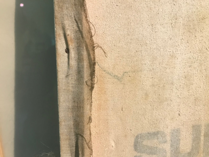May include: Close-up of a beige canvas with a frayed edge. The canvas has a brown fabric border and a small hole in the center. The canvas is slightly textured and has a few faint marks.