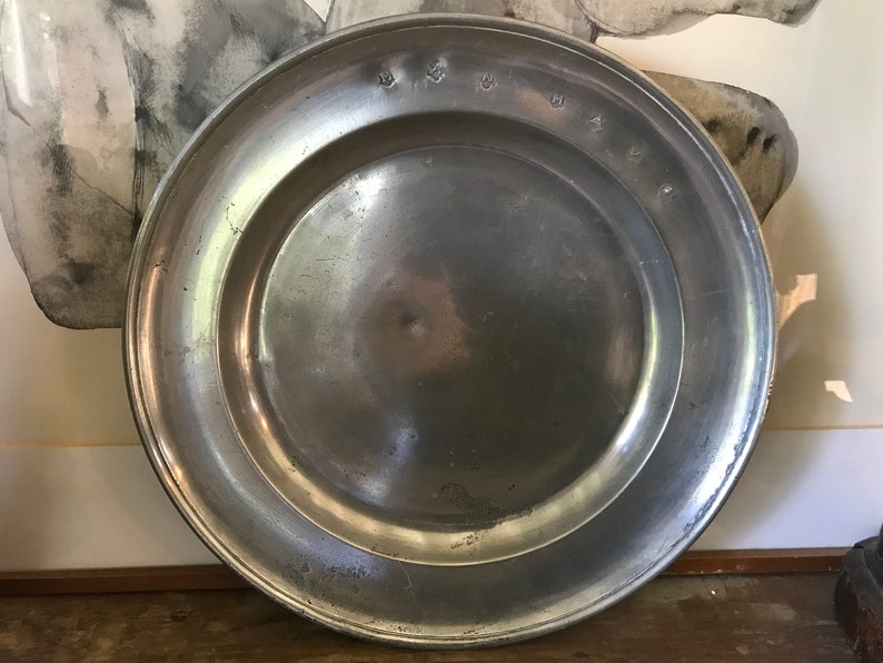 May include: A large, round, silver pewter plate with a raised rim. The plate has a smooth, shiny surface and is slightly tarnished. The plate is marked with the letters "W & W" on the rim.