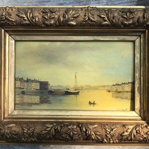 Op de afbeelding: Een ingelijst olieverfschilderij dat een zeegezicht met een zeilboot en gebouwen langs de waterkant toont. De lucht is zachtgeel en blauw, en het water is kalm en reflecterend blauw. Het schilderij is in een gouden lijst met sierlijke details.