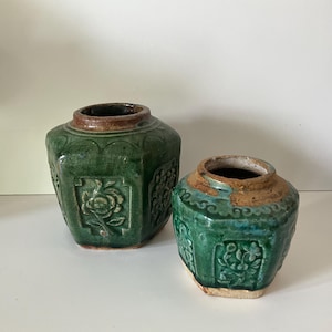 Peut inclure: Deux pots en céramique verte antiques avec des motifs floraux en relief. Les pots présentent un aspect vieilli, avec des fissures visibles et des variations dans l'émail. Les bords sont de couleur marron contrastante.