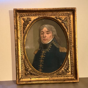 Op de afbeelding: Een ingelijst portret van een man in militair uniform. De man heeft grijs haar en draagt een blauw jasje met gouden knopen en een witte kraag. Het portret is ingelijst in een gouden lijst met sierlijke details.