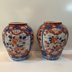Puede incluir: Dos jarrones de cerámica a juego con una forma ancha y redondeada. Los jarrones presentan un fondo blanco con intrincados diseños florales en naranja y azul. Los bordes son de color rojo intenso, y la estética general recuerda a la cerámica asiática tradicional.