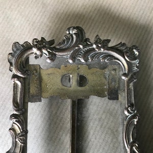 May include: An ornate silver-toned picture frame with a glass front and a decorative top. The frame features a detailed design with scrolls and floral motifs.