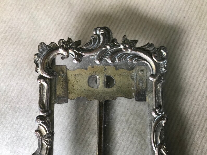 May include: An ornate silver-toned picture frame with a glass front and a decorative top. The frame features a detailed design with scrolls and floral motifs.