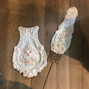 May include: Two white and pink decorative wall plaques with intricate floral designs. The plaques are made of plaster or ceramic and have a vintage or antique aesthetic.
