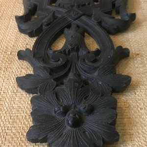 May include: A black, intricately carved wooden ornament with a floral design. The ornament has a symmetrical design with a central flower and swirling patterns.