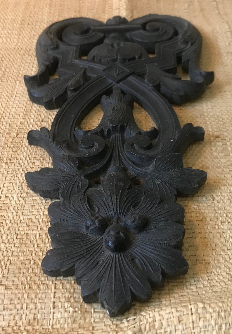 May include: A black, intricately carved wooden ornament with a floral design. The ornament has a symmetrical design with a central flower and swirling patterns.