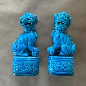 Puede incluir: Dos estatuas a juego de perros Foo de cerámica turquesa. Cada estatua está sentada sobre una base decorativa. Los perros Foo tienen detalles, incluyendo bocas abiertas, y son de un tono azul vibrante.