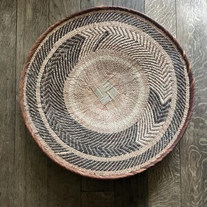 May include: A large, round, woven basket with a brown and white geometric pattern. The basket is made of natural materials and has a slightly raised center.