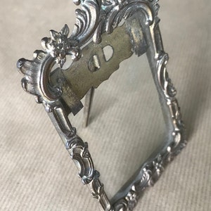 May include: A silver-toned metal picture frame with an ornate design. The frame has a stand on the back and a glass front.