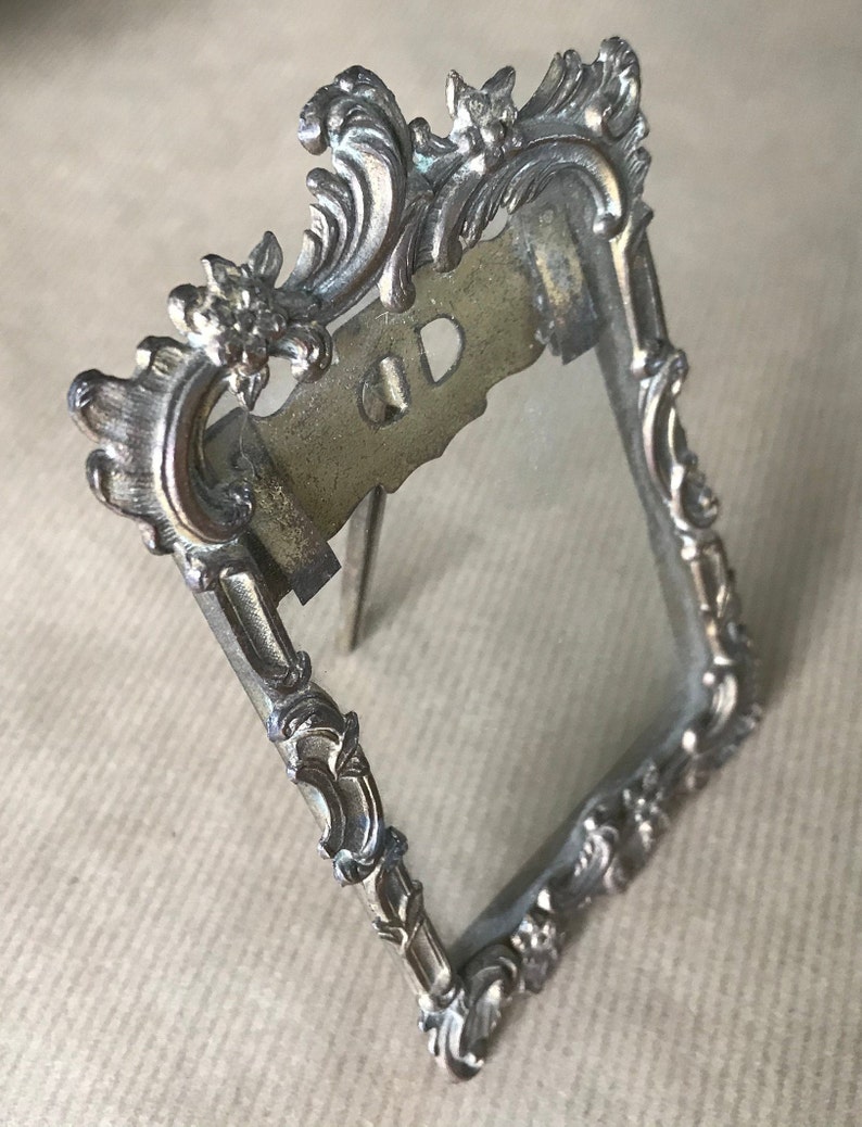 May include: A silver-toned metal picture frame with an ornate design. The frame has a stand on the back and a glass front.