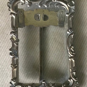 May include: A vintage silver-toned picture frame with an ornate, scrolled design. The frame has a glass front and a metal back.