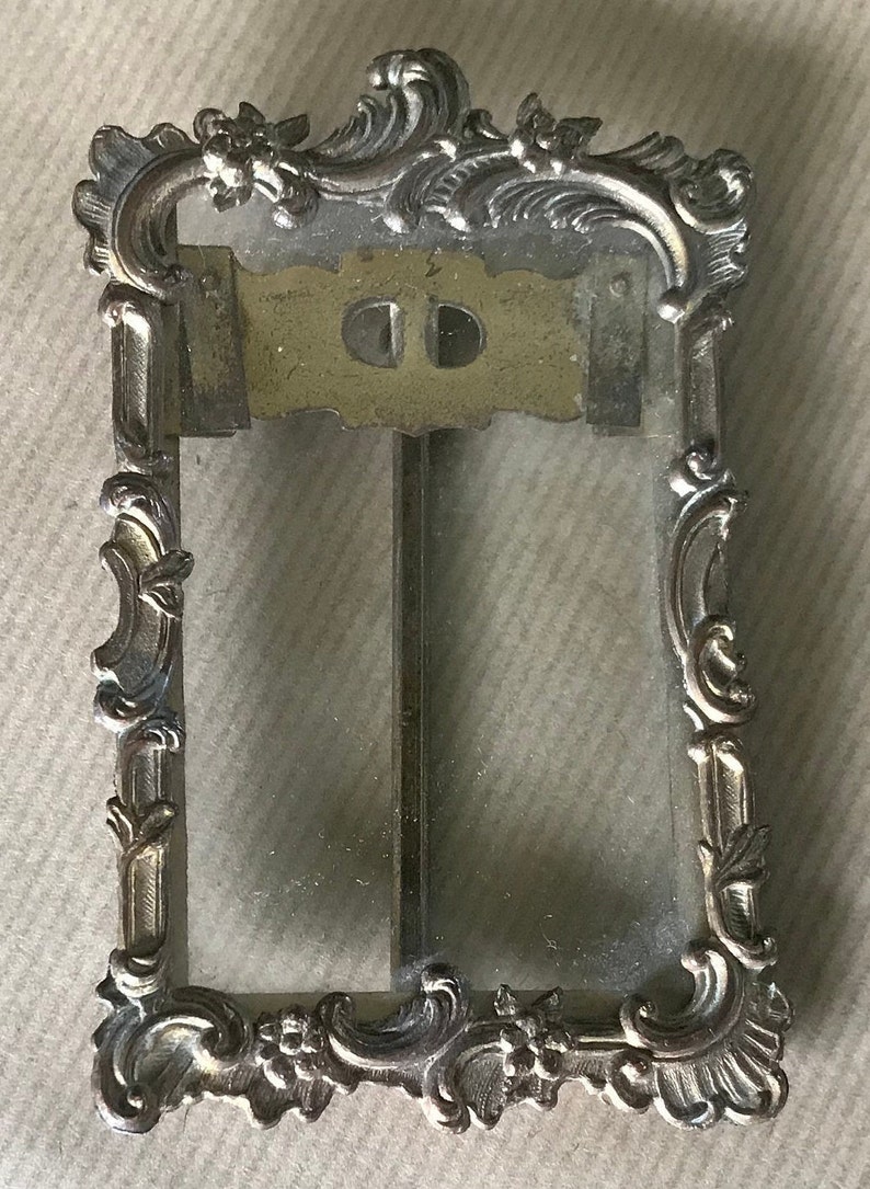 May include: A vintage silver-toned picture frame with an ornate, scrolled design. The frame has a glass front and a metal back.