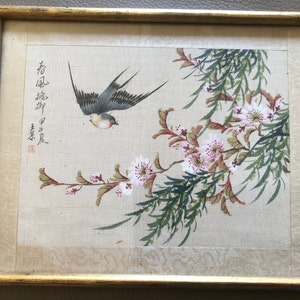 May include: A framed painting of a bird in flight with pink flowers and green leaves. The painting is on a beige background and has Chinese characters in the top left corner.