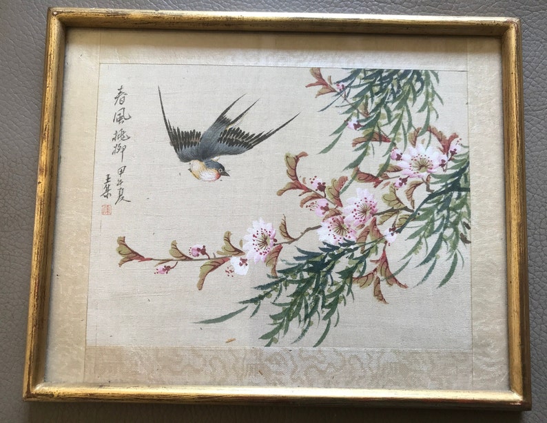 May include: A framed painting of a bird in flight with pink flowers and green leaves. The painting is on a beige background and has Chinese characters in the top left corner.