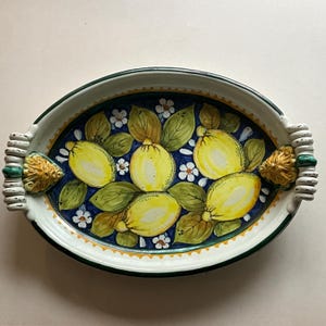 Puede incluir: Plato de servir ovalado de cerámica con asas, con un diseño pintado a mano de limones, hojas y flores blancas sobre un fondo azul. El plato tiene un borde blanco con un ribete verde y dorado.
