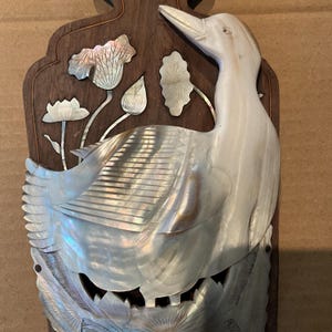 Könnte beinhalten: Eine dunkelbraune Wandplatte aus Holz mit einer schillernden Perlmutt-Einlage, die einen großen Vogel inmitten von Lotusblumen und Blättern zeigt. Die Einlage hat eine schimmernde Oberfläche mit detaillierten Gravuren auf dem Vogel.