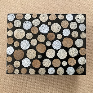 Puede incluir: Una caja negra con un patrón decorativo de círculos blancos y marrones. Los círculos están hechos de un material que parece madera y tienen una textura agrietada.