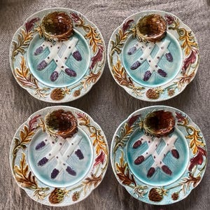 May include: Set of four decorative ceramic plates. Each plate features a light blue center with a crossed asparagus design and a small brown bowl. The rims are adorned with a detailed leaf and floral pattern in shades of brown, gold, and burgundy.