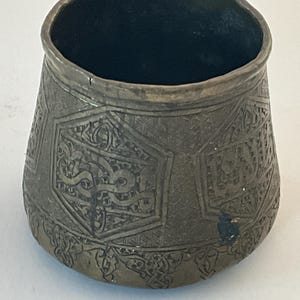 May include: A small, round, brass bowl with intricate engraved designs. The bowl has a hexagonal pattern with Arabic script.