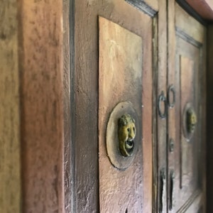 Op de afbeelding: Close-up van een houten kast met twee deuren. De deuren hebben sierlijke messing beslag en zijn geschilderd in een rustieke bruine kleur.