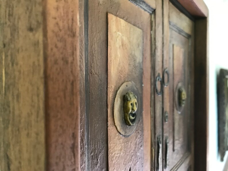 Op de afbeelding: Close-up van een houten kast met twee deuren. De deuren hebben sierlijke messing beslag en zijn geschilderd in een rustieke bruine kleur.