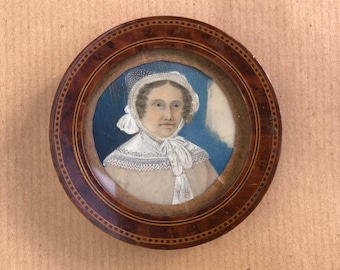 May include: A framed miniature portrait of a woman wearing a white lace bonnet and a white lace collar. The portrait is painted on ivory and set in a wooden frame with a dark brown finish.