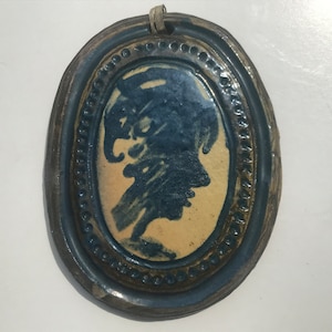 May include: An oval ceramic ornament with a blue and white glaze. The ornament features a blue silhouette of a person's head with a hat on. The ornament has a brown rope for hanging.