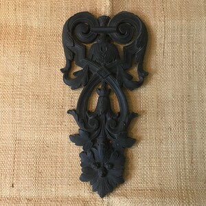 May include: A black, ornate, decorative wall hanging with a floral design. The design is intricate and features a central motif with a diamond shape and a flower at the bottom.