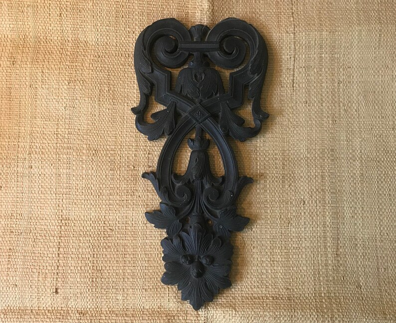 May include: A black, ornate, decorative wall hanging with a floral design. The design is intricate and features a central motif with a diamond shape and a flower at the bottom.