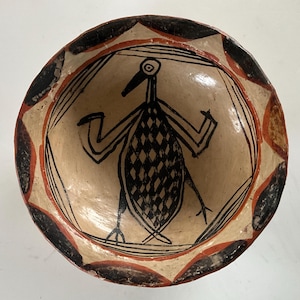 Op de afbeelding: Een ronde, beige keramische kom met een handgeschilderd vogelontwerp. De vogel is zwart met een ruitpatroon op zijn lichaam. De rand heeft een herhalend patroon van zwarte en rode vormen.