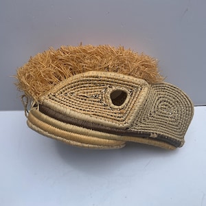May include: A woven mask shaped like a bird's head, made from natural straw and reeds. It has a circular eye hole and a textured, straw-like crest. The beak and head are intricately woven, showcasing detailed craftsmanship.