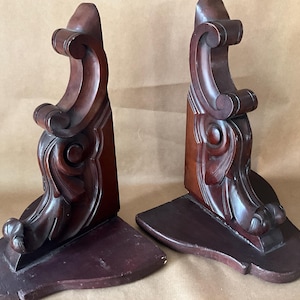Puede incluir: Par de sujetalibros antiguos de madera. Cada sujetalibros presenta un diseño ornamentado de color marrón oscuro con detalles tallados y enrollados. La base es rectangular y el estilo general recuerda a los muebles antiguos.