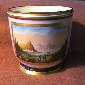 Puede incluir: Un jarrón de porcelana rosa con un borde dorado y una escena pintada de un lago con montañas y un velero. El jarrón tiene una banda marrón alrededor de la parte superior e inferior.