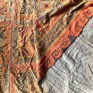 Op de afbeelding: Een gedetailleerd, patroonrijk textiel in tinten oranje, bruin en paars. De stof heeft geometrische en bloemmotieven, met een geschulpte rand in een donkerdere oranje kleur. Het textiel is waarschijnlijk een decoratief stuk, zoals een sprei of wandkleed.