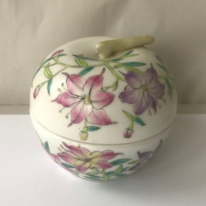 Puede incluir: Un tarro de porcelana blanca con tapa en forma de manzana con un tallo verde. El tarro está decorado con flores rosas, moradas y amarillas y hojas verdes.
