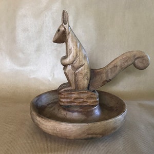 Puede incluir: Un cascanueces de madera con forma de canguro. El canguro está de pie sobre una base de madera con un cuenco para contener las nueces. La cola del canguro es la palanca para romper las nueces.