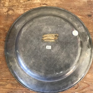 May include: A silver-colored metal plate with a raised rim. The plate has a label on the back that reads "Museum Purchase 1844. Gift of Mrs. E. B. 375."