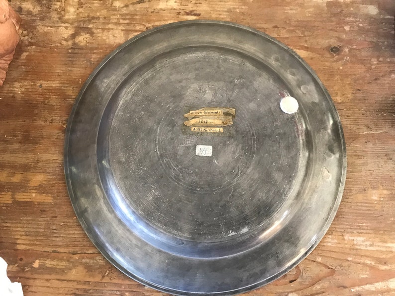 May include: A silver-colored metal plate with a raised rim. The plate has a label on the back that reads "Museum Purchase 1844. Gift of Mrs. E. B. 375."