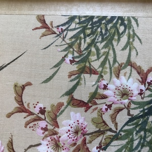 May include: A close-up of a delicate floral painting on a beige fabric background. The painting features pink and white flowers with green leaves and branches. The flowers have a delicate, watercolor-like appearance.