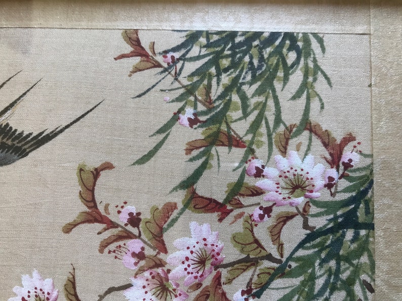 May include: A close-up of a delicate floral painting on a beige fabric background. The painting features pink and white flowers with green leaves and branches. The flowers have a delicate, watercolor-like appearance.