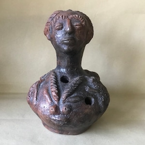 Può includere: Una scultura in terracotta di una testa e spalle di una persona con un viso dettagliato e un design decorativo sul petto. La scultura presenta diversi fori nella zona del petto.