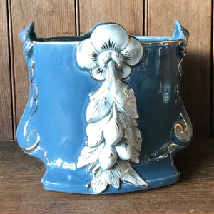 May include: A blue ceramic planter with white and gold floral accents. The planter has a unique, ornate design with a flower and leaves on the front.
