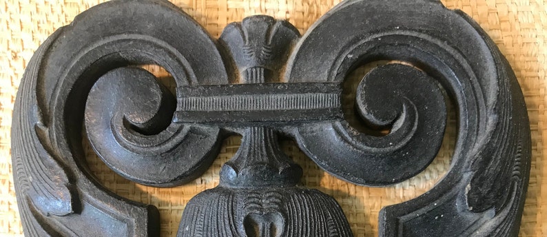 May include: A black wooden decorative piece with intricate carvings in the shape of a heart. The piece has a central design with a floral motif and two symmetrical scrolls on either side.