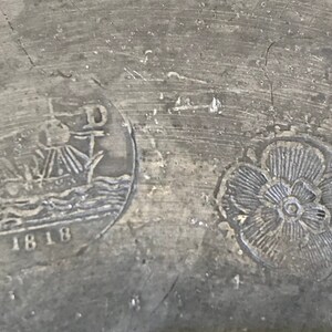 May include: Close-up of a pewter plate with two embossed designs. The left design features a ship with the date 1818 below it. The right design is a stylized flower.